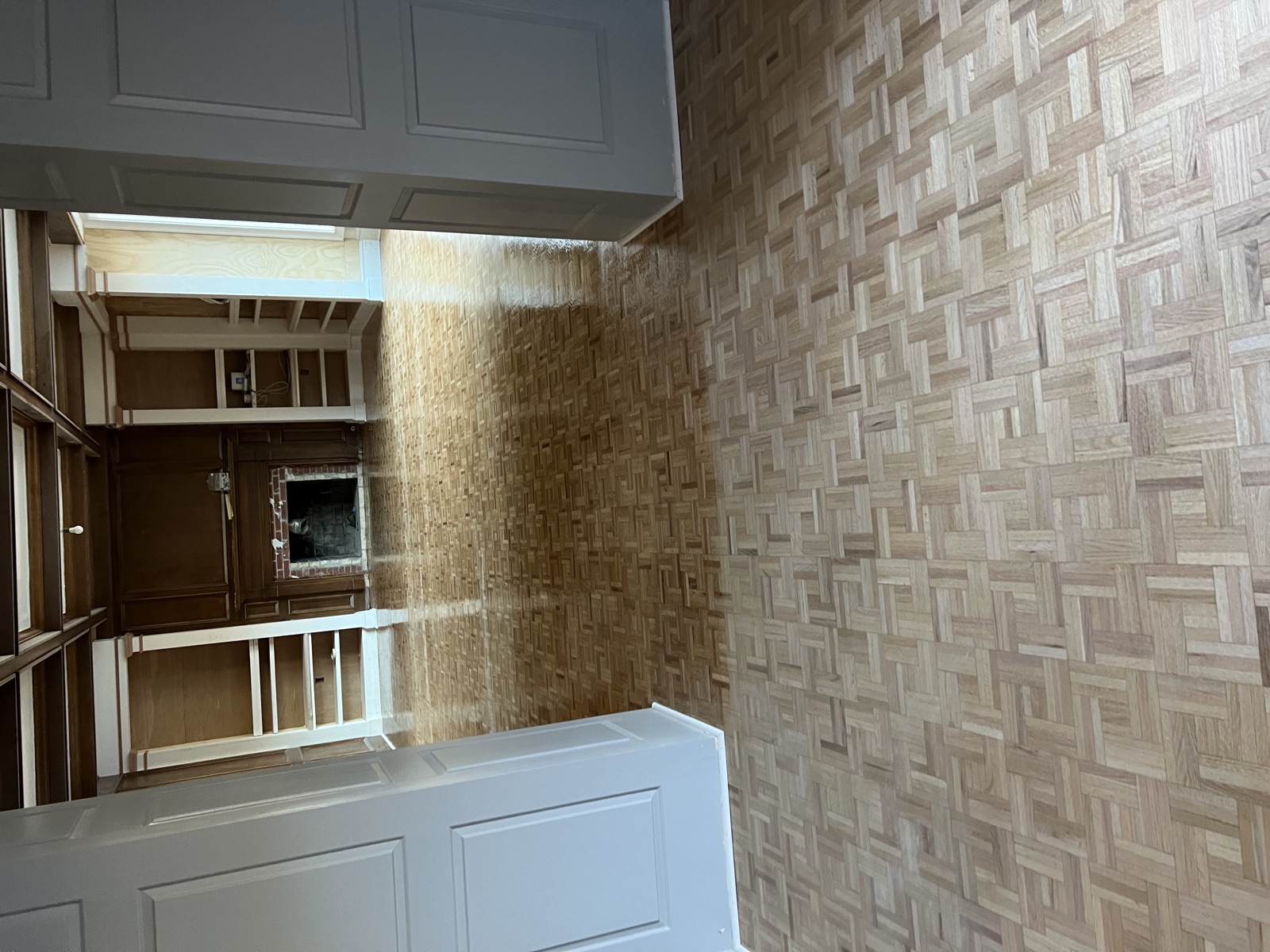 Basket-weave parquet hardwood flooring viewed through open doorway into formal room with wood paneling, coffered ceiling, and brick fireplace