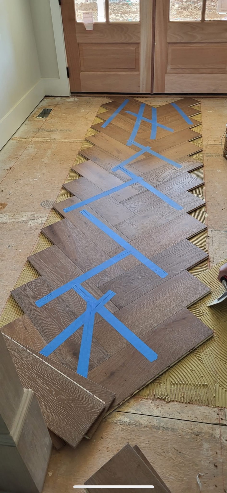 Herringbone hardwood flooring being installed in foyer entryway with blue painter's tape holding planks in place